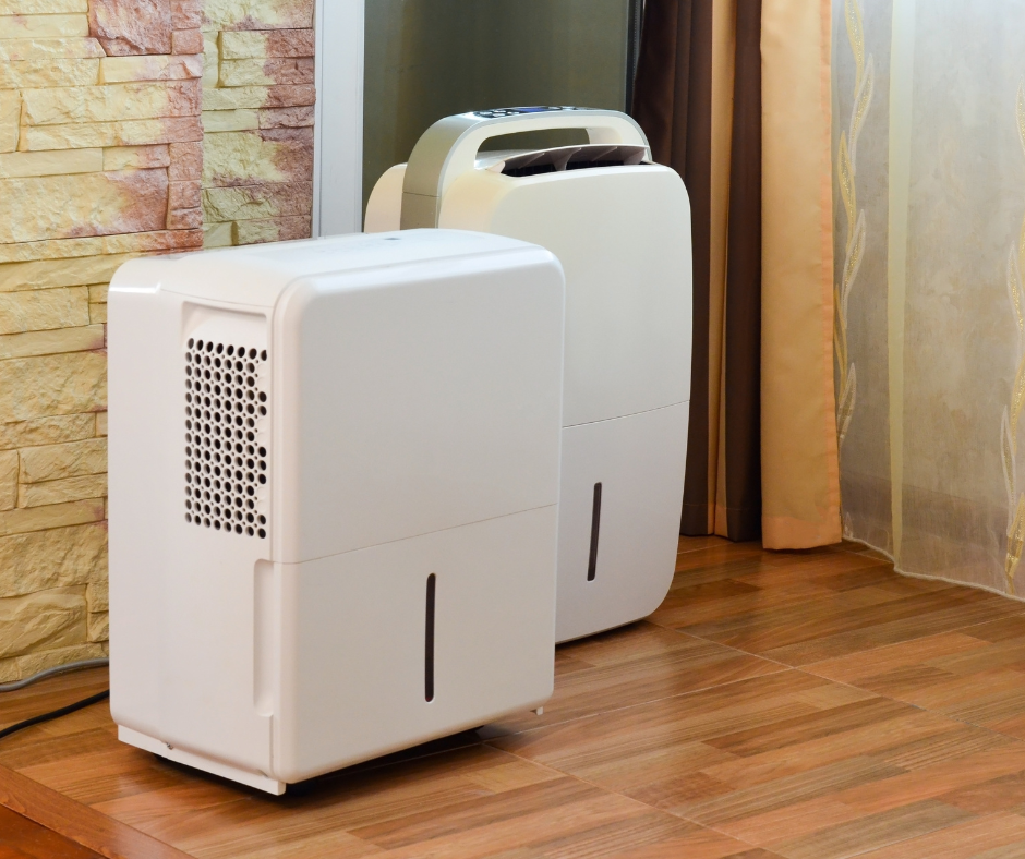 Two white dehumidifiers placed on a wooden floor inside a home, positioned near a wall and curtains to reduce indoor moisture.