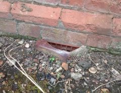 A rusty metal vent is set into the base of a red brick wall, surrounded by gravel, small rocks, soil, and dry twigs.