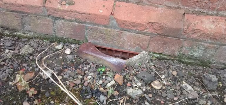 A rusty metal vent is set into the base of a red brick wall, surrounded by gravel, small rocks, soil, and dry twigs.