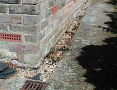 A close-up view of a building’s brick corner, with an external wall vent, gravel along the foundation, and a concrete path running beside the wall.