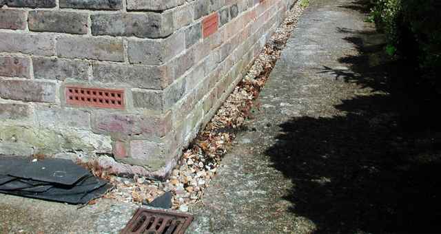A close-up view of a building’s brick corner, with an external wall vent, gravel along the foundation, and a concrete path running beside the wall.